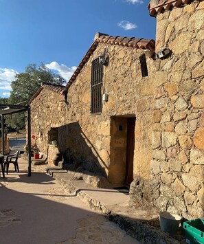 Un edificio de piedra con techo de tejas, una puerta y una ventana con barrotes. Hay una silla y una mesa afuera, y un caja verde cerca.