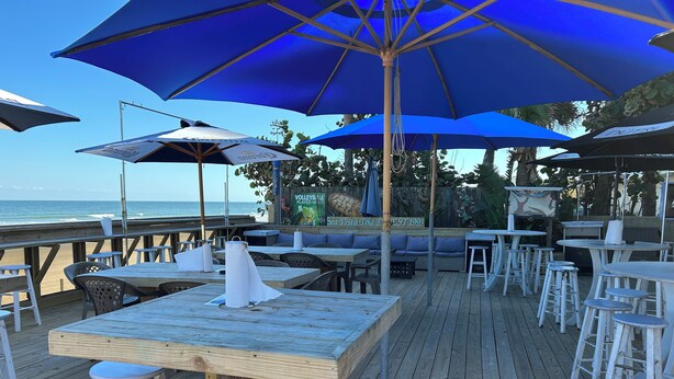 Eine Holzterrasse mit Tischen und Stühlen, blauen Sonnenschirmen und Blick auf das Meer.