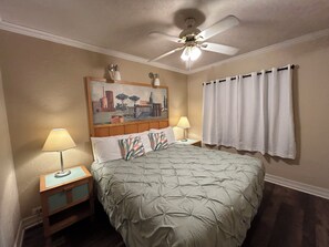 A bedroom with a bed, two bedside tables, a ceiling fan, a painting, and a window with curtains. - Sea Vista Resort And Tiki Bar (New Smyrna Beach)
