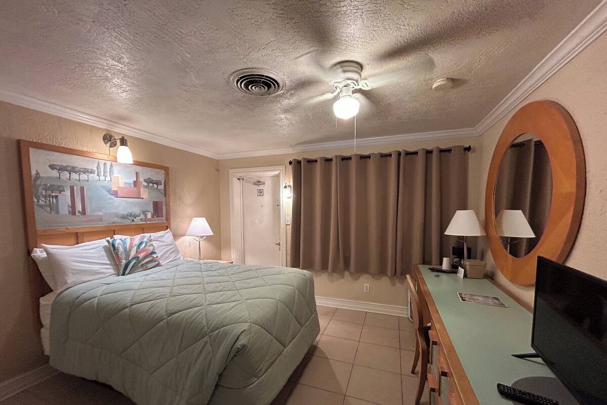 Ein Hotelzimmer mit einem Bett, einem Schreibtisch mit Computer, einem Fernseher, einem Deckenventilator und einem Gemälde an der Wand.