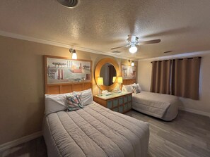 A hotel room with two beds, a ceiling fan, a painting on the wall, a round mirror, and a window with curtains. - Sea Vista Resort And Tiki Bar (New Smyrna Beach)
