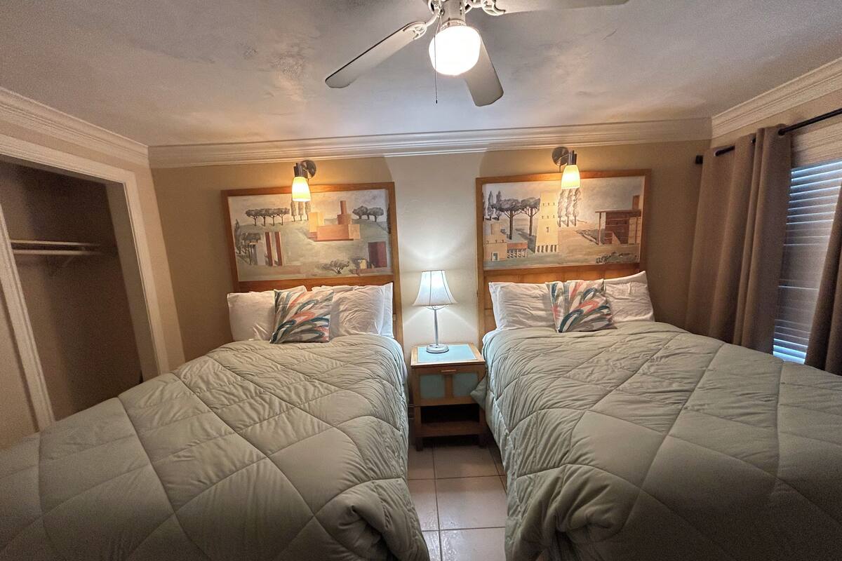 Ein Hotelzimmer mit zwei Betten, einem Deckenventilator, Kunstwerken an den Wänden und einem Nachttisch mit Lampe.