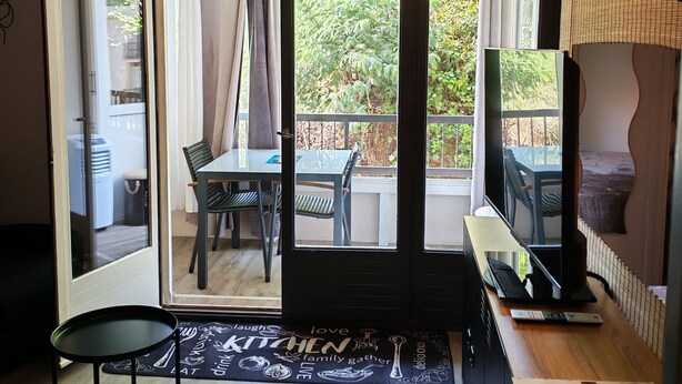 Une pièce dotée d’une porte-fenêtre coulissante donnant sur un balcon extérieur, d’un ventilateur de plafond, d’une table à manger avec des chaises et d’un téléviseur posé sur un meuble.