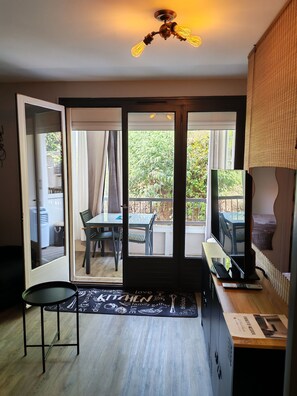 A room with a sliding glass door leading to an outdoor balcony, a ceiling fan, a dining table with chairs, and a television on a stand.