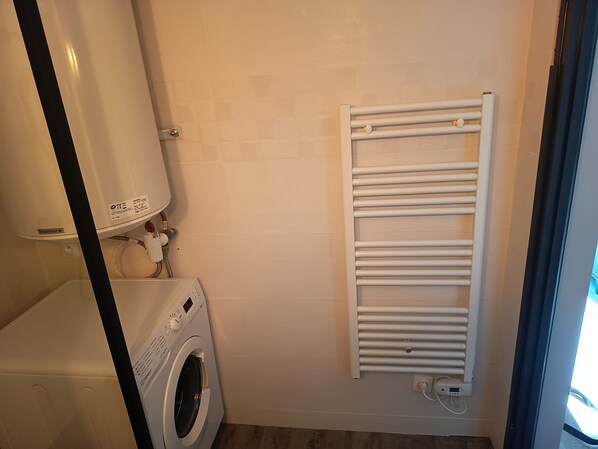 A compact laundry room with a front-loading washing machine, a white towel rack, and a wall-mounted heater.