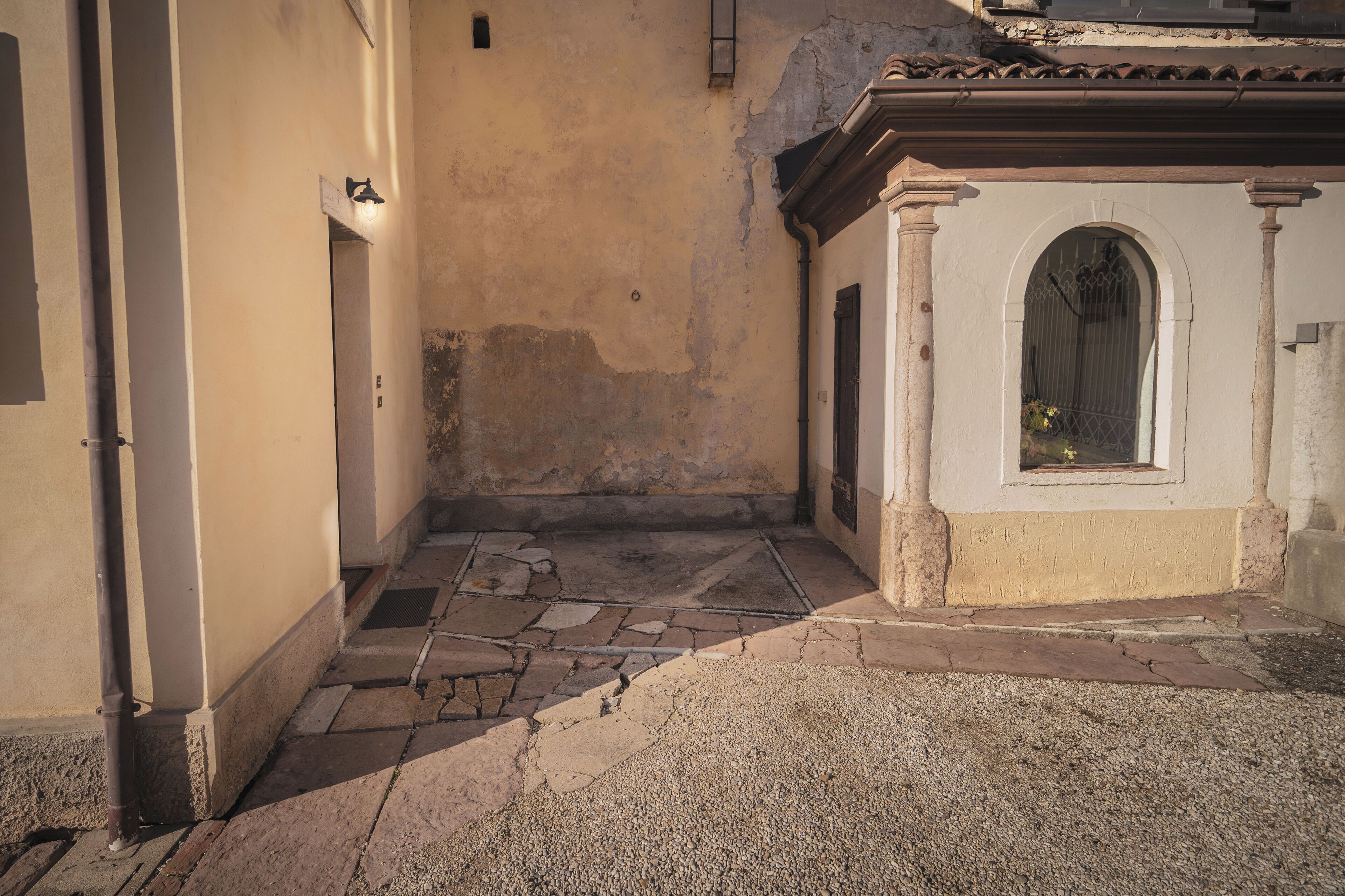 A narrow alleyway with a stone-paved path leading to a building with a small arched window and a door.