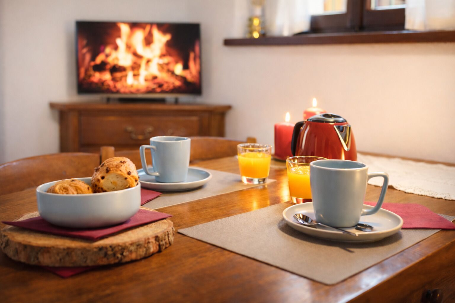 Une table en bois dressée pour le petit-déjeuner, avec un bol de viennoiseries, deux tasses, deux verres de jus d’orange et une cafetière rouge.