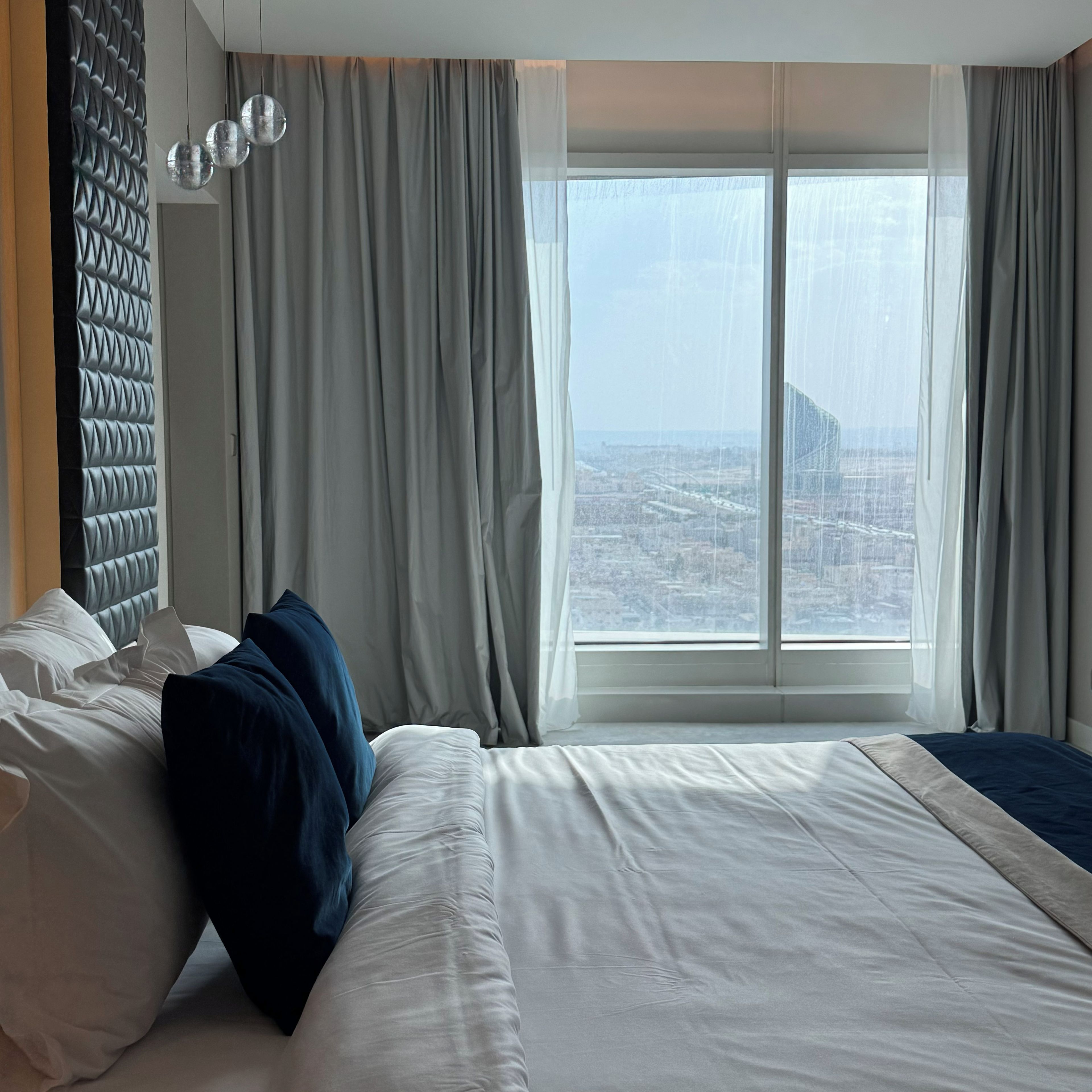 Habitación de hotel con una cama grande, vista de la ciudad a través de una ventana y un cabecero moderno.