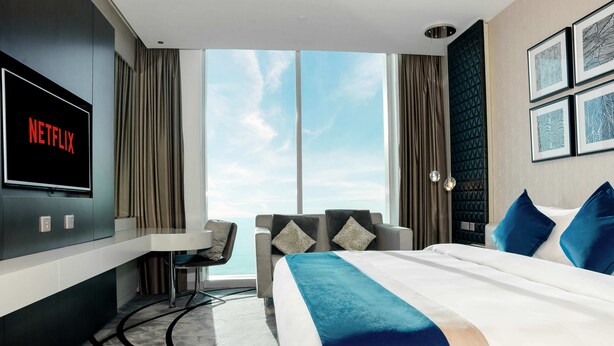 A hotel room with a large bed, a desk, a chair, a TV displaying 'NETFLIX', and a view of the sky through large windows. - Afaq Luxury - Al-Olya (Riyadh)