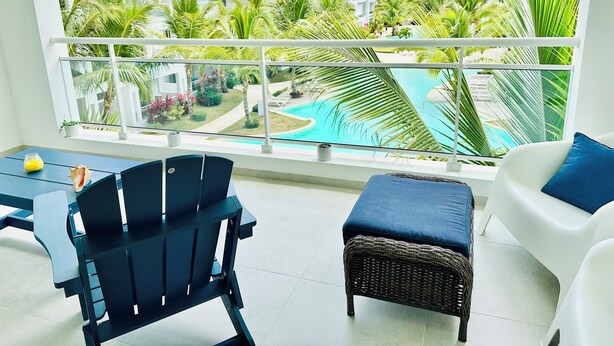 Ein Balkon mit Blick auf ein Schwimmbad und Palmen, ein Esstisch mit Stühlen sowie ein Korbsessel.