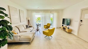 Ein modernes Wohnzimmer mit einer Couch, zwei gelben Sesseln, einem Fernseher und einem Balkon mit Vorhängen.