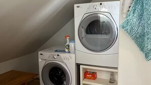 Ein Waschbereich mit zwei übereinander gestapelten Waschmaschinen, einer kleinen Holzbank und einem Regal mit einer Kiste und einer Sprühflasche.