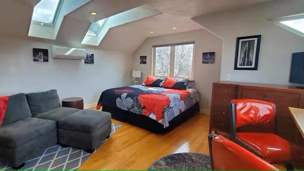 Ein modernes Schlafzimmer mit einer Schlafgalerie, einem Bett mit floralem Überwurf, einem grauen Sofa, einem roten Sessel und einem Fernseher auf einem Holzkabinett.