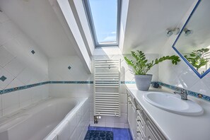 Ein modernes Badezimmer mit Oberlicht, weißen Fliesen, einer Badewanne, einem Waschbecken und einer Topfpflanze.