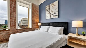 A hotel room with a large bed, a city view through a window, a brick wall, and a modern art piece on the wall. - Dallas Skyline Brick Loft, Sleeps 7 - Free Valet & Pool (Dallas)