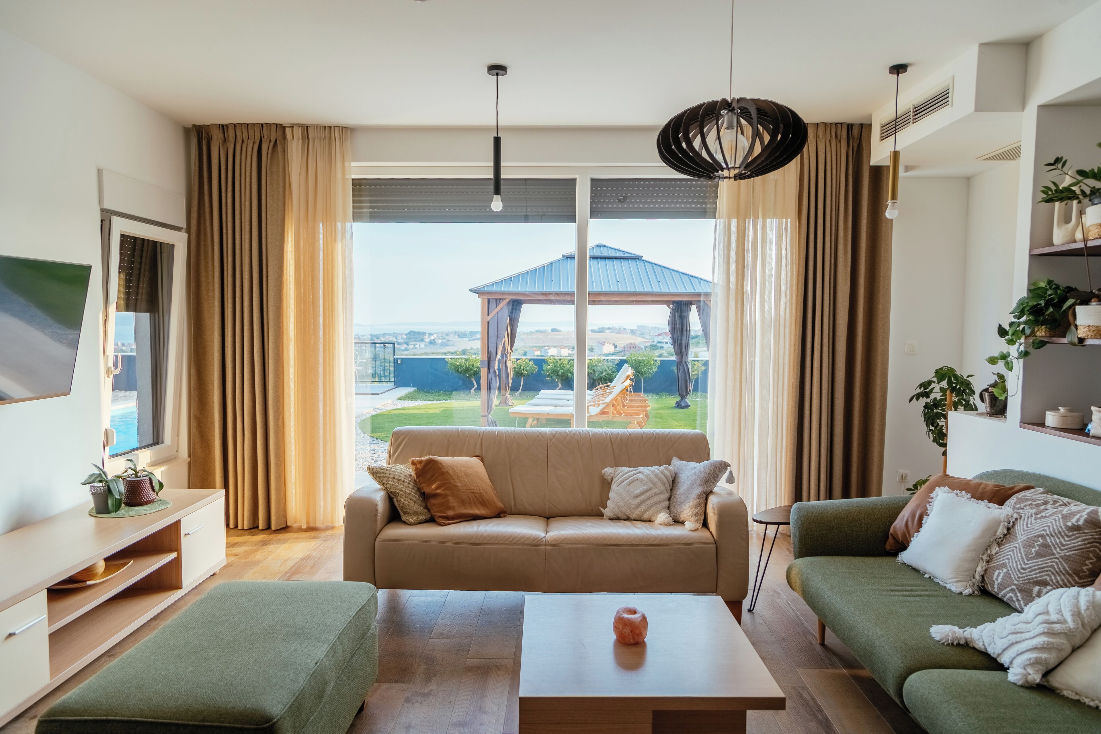 Un salon moderne avec un canapé, une table basse et un téléviseur. Il y a une grande fenêtre avec des rideaux, un luminaire au plafond et une vue sur un jardin et un kiosque.