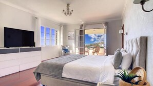 Quarto com cama grande, televisão na parede, varanda com cadeiras e vista do céu.
