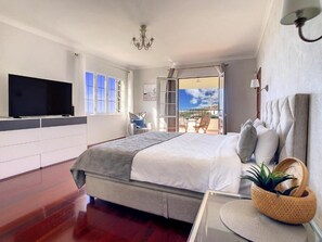 A bedroom with a large bed, a TV on the wall, a balcony with chairs, and a view of the sky. - Villa Baradaye by Atlantic Holiday (Estreito Calheta)