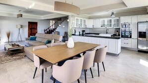 Ampla sala de estar com mesa e cadeiras de jantar, sofá e cozinha com armários brancos e eletrodomésticos modernos.