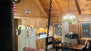 Ein gemütliches Blockhaus-Interieur mit Holzwänden und hohen Decken, mit einer Küche inklusive Kühlschrank, Herd und Mikrowelle, einem Essbereich mit Tisch und Stühlen sowie einem großen Fenster mit Vorhängen.
