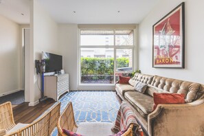 Ein Wohnzimmer mit einer Couch, einem Fernseher, einem gemusterten Teppich und einem großen Fenster mit Blick auf Grünflächen.