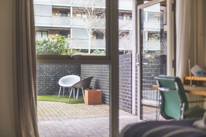 Blick durch eine Glastür in einen Innenhof mit einer Ziegelwand, einem weißen Stuhl und einem grünen Stuhl.