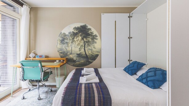 Een slaapkamer met een bed, een bureau, een stoel, een kledingkast en een groot rond schilderij aan de muur.