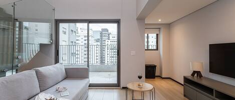 Een moderne woonkamer met een bank, een klein rond tafeltje, een televisie en een balkon met uitzicht op de stad.