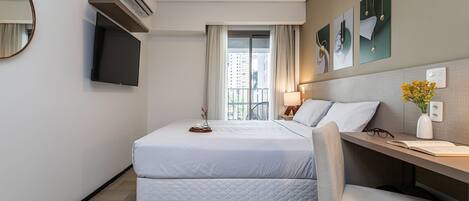 Habitación de hotel con cama, escritorio, silla, televisor, unidad de aire acondicionado, ventana con cortinas, obra de arte y un jarrón con flores.