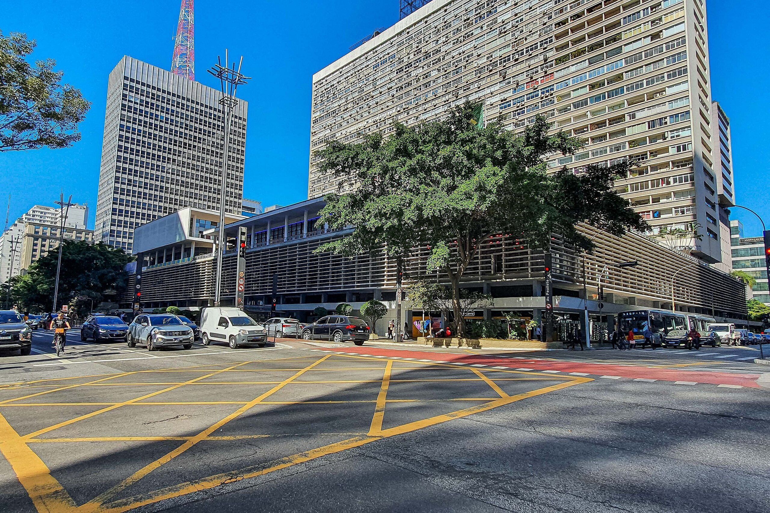 A city intersection with tall buildings, a tree-lined street, and a clear blue sky.