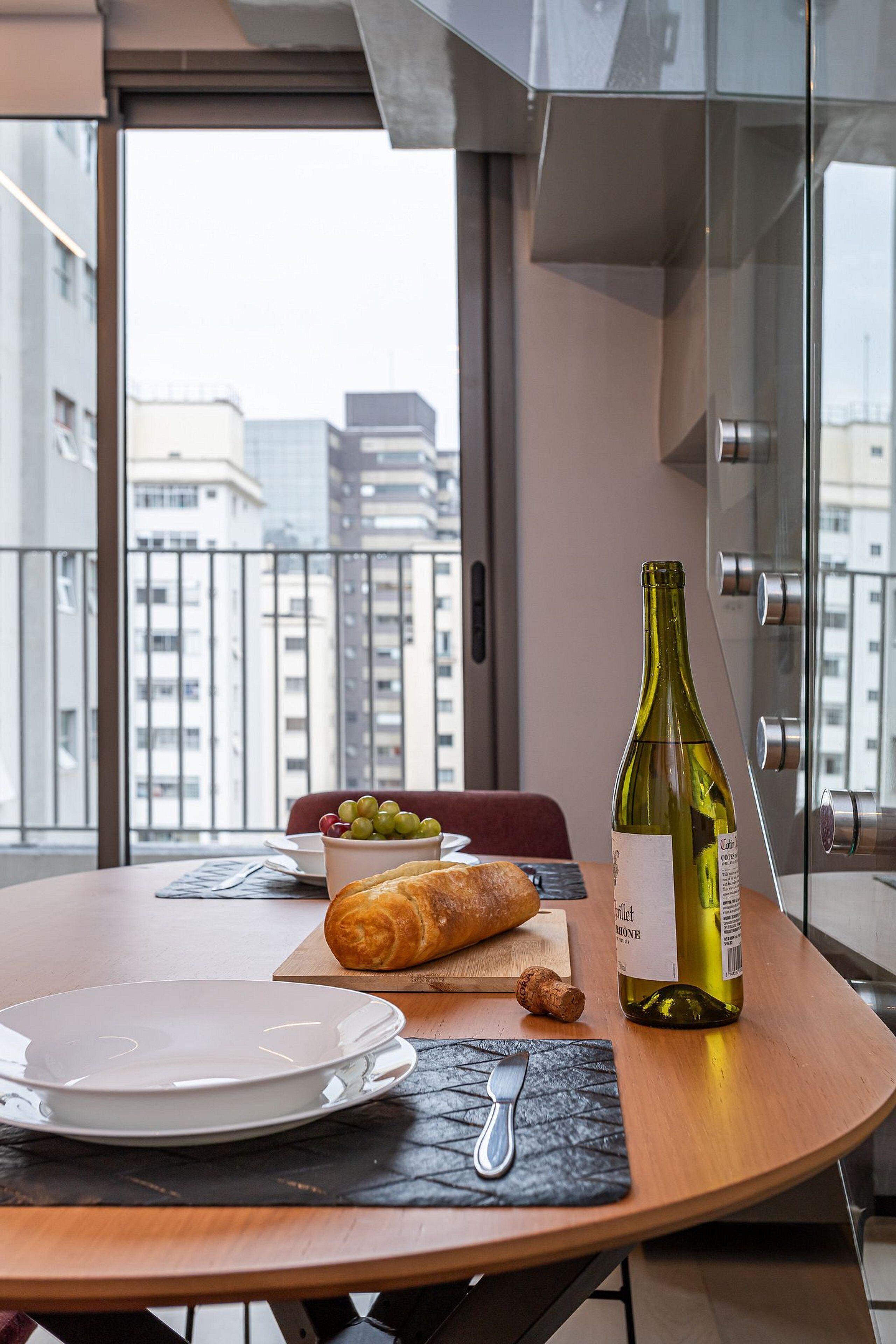 A dining table with a bottle of wine, a croissant, and a bowl of grapes on a balcony with a city view.