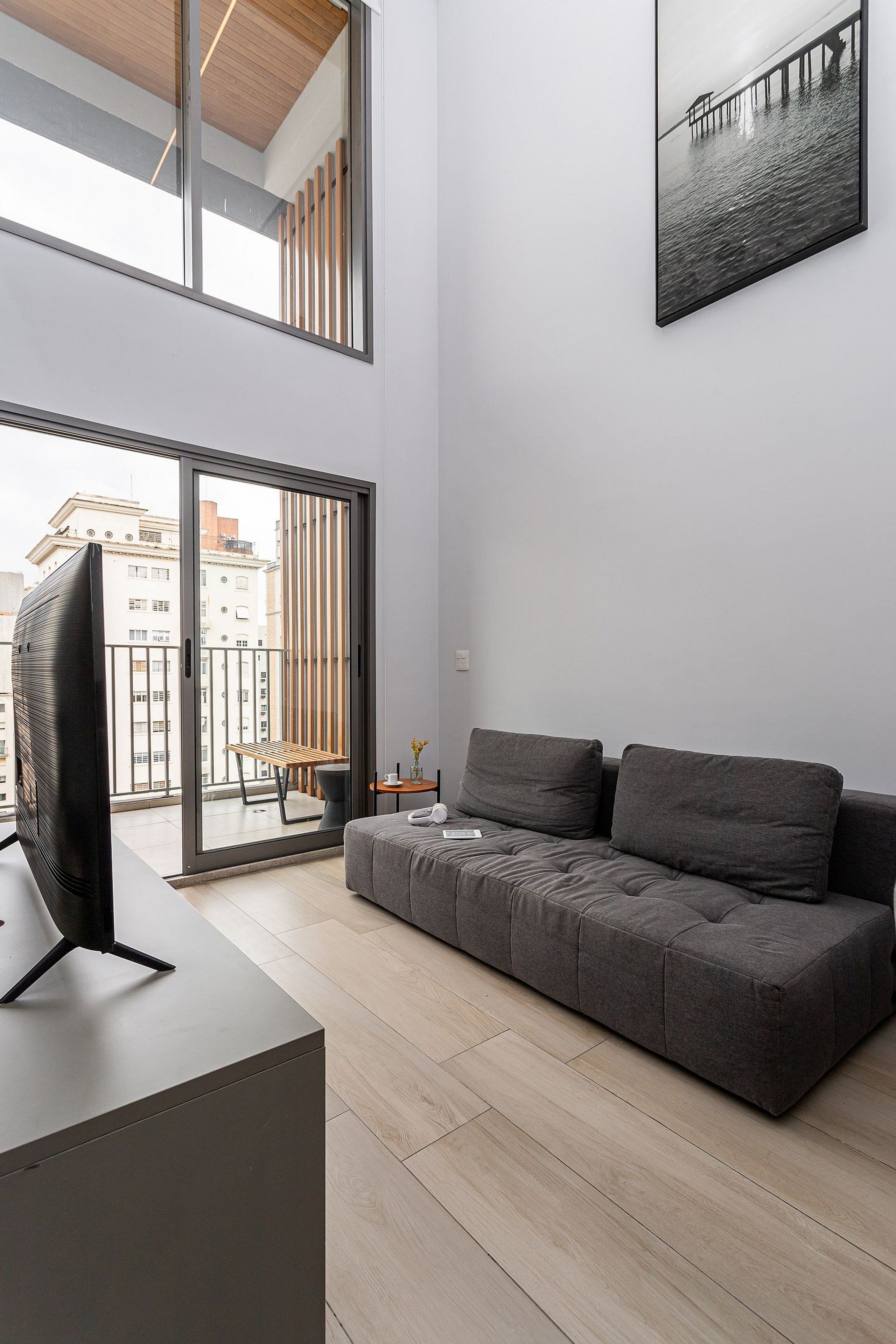 A modern living room with a sofa, a flat-screen TV, and a balcony with a view of buildings.