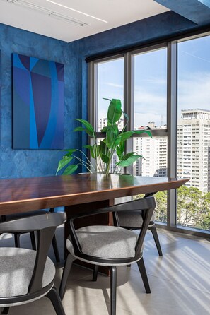 A modern room with a large window, a wooden table, two grey chairs, a green plant, and a blue abstract painting on the wall. - Pivô Jardins by Viva - Apartamentos Novos e Modernos (São Paulo)