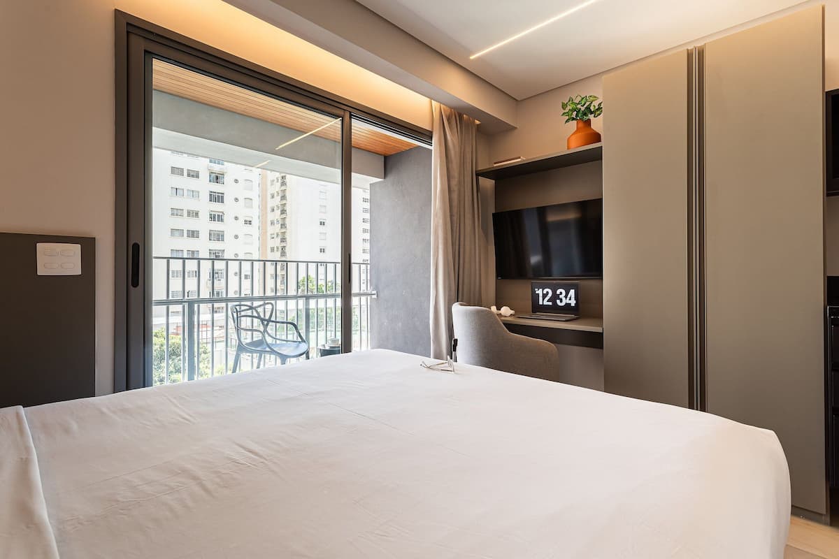 Habitación de hotel con una cama grande, televisor, silla y balcón con vista a edificios.