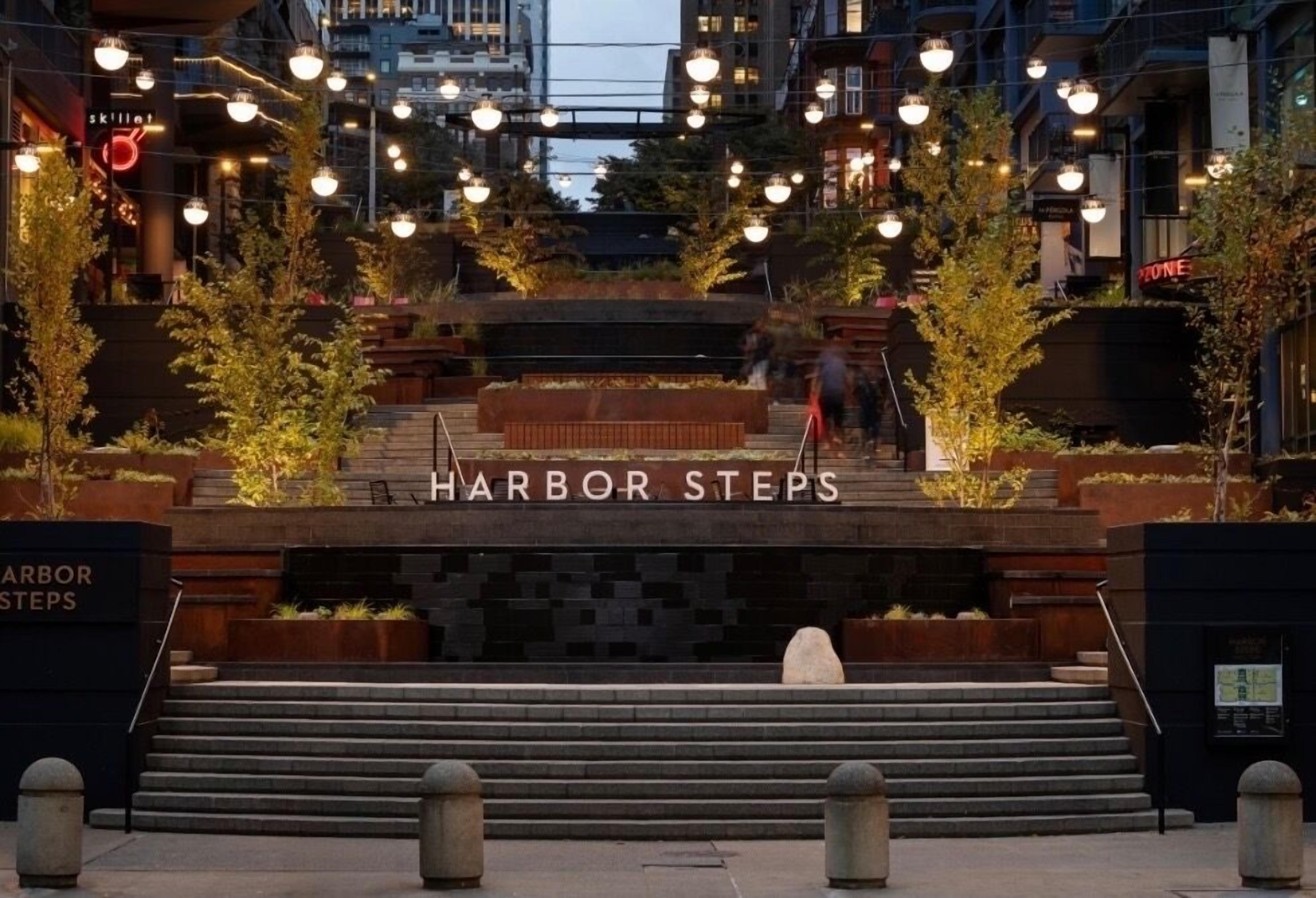 Een openbaar gebied met trappen die leiden naar een bord waarop 'Harbor Steps' staat, omringd door bomen en lantaarns.
