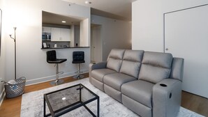 Una sala moderna con un sofá gris, una mesa de centro de vidrio y una cocina visible a través de un arco.