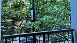 Un balcón con vista a un edificio de varios pisos, una calle con coches aparcados y exuberantes árboles verdes.