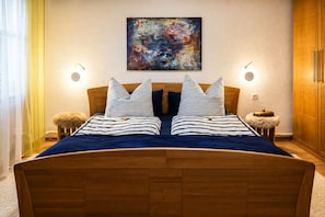 A bed with a wooden headboard, two bedside tables with lamps, a fluffy stool, and a painting on the wall. - New - Stylish granny apartment with modern living comfort on 65 sqm (Oberschönau)