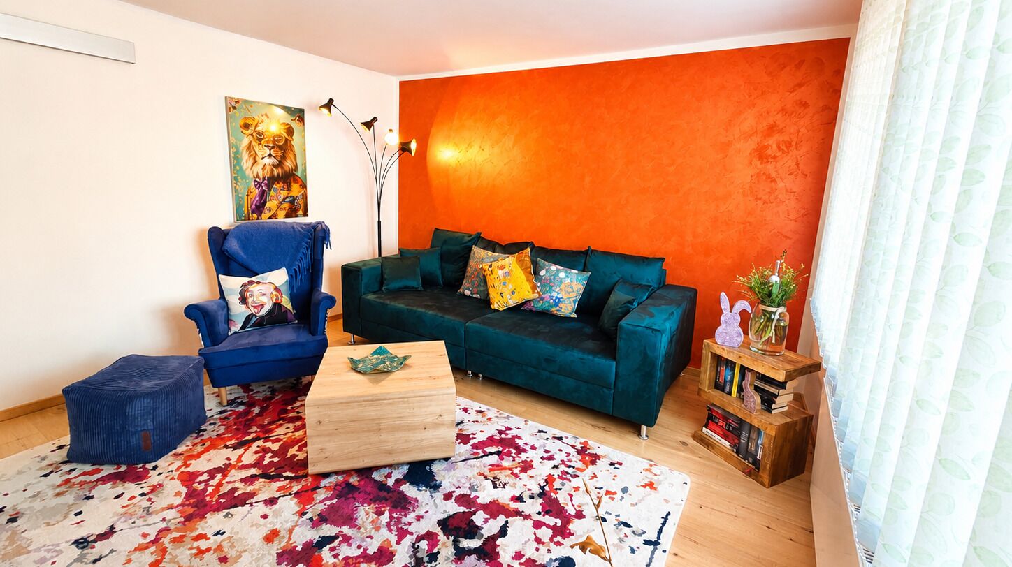 Een woonkamer met een blauwgroene bank, een blauwe fauteuil, een kleurrijk vloerkleed, en een boekenkast met boeken en een plant.