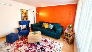 Een woonkamer met een blauwgroene bank, een blauwe fauteuil, een kleurrijk vloerkleed, en een boekenkast met boeken en een plant.