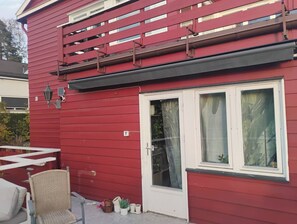 Un bâtiment rouge avec une porte et des fenêtres blanches, un petit espace extérieur aménagé avec une chaise, et une rambarde en métal.