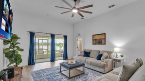 A spacious living room with a large window, a ceiling fan, a sofa, a coffee table, and a TV mounted on the wall. - Dinner Lake Home | Sunset Views | Extended Stay | 4BR, Sleeps 8, Near Legoland (Lake Wales)