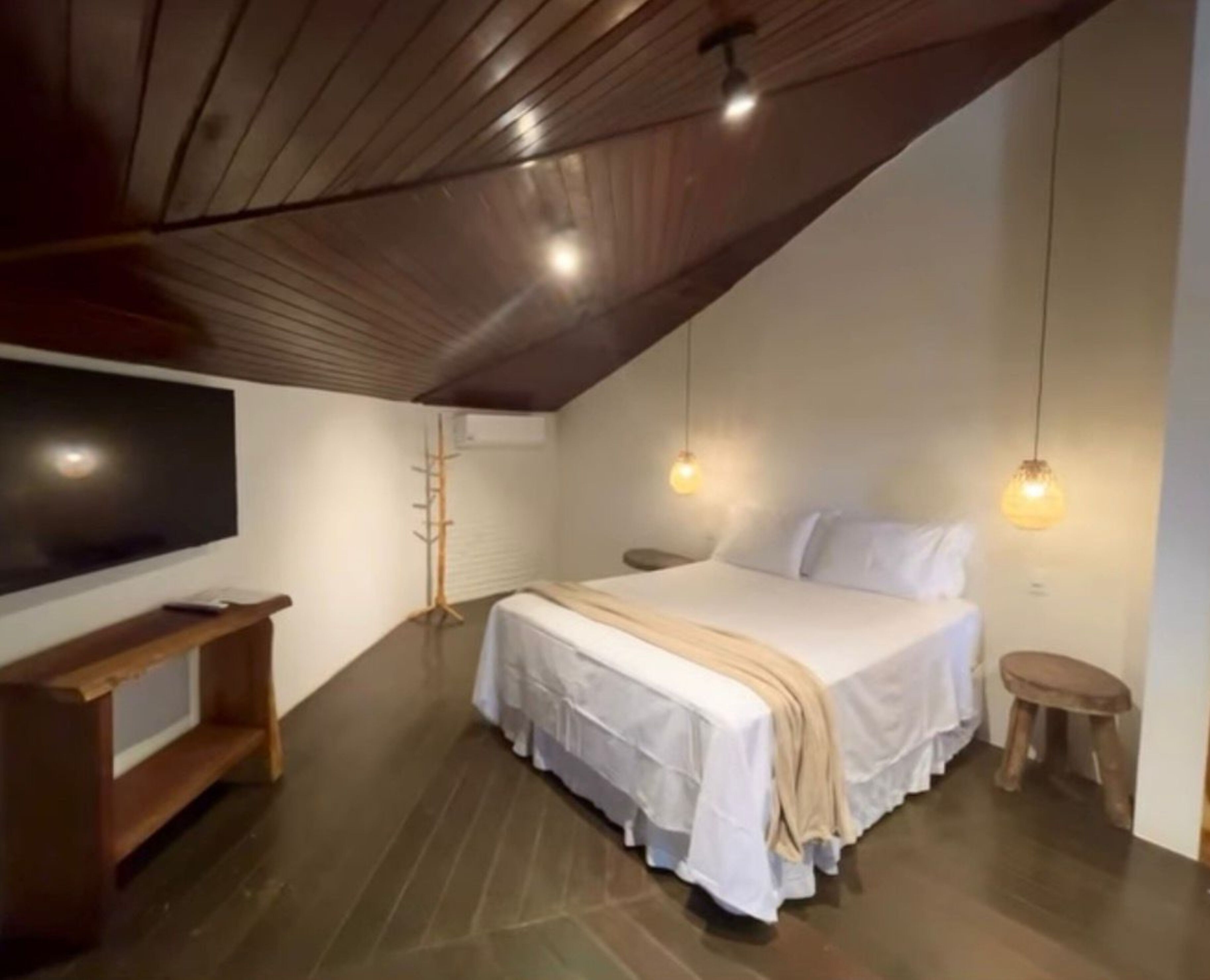 A bedroom with a sloped ceiling, a bed with white bedding, a wooden bench, a TV, and pendant lights.