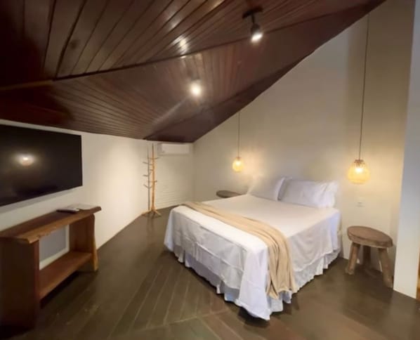 A bedroom with a sloped ceiling, a bed with white bedding, a wooden bench, a TV, and pendant lights. - Pousada Vila Margarida (Chapada dos Guimarães)