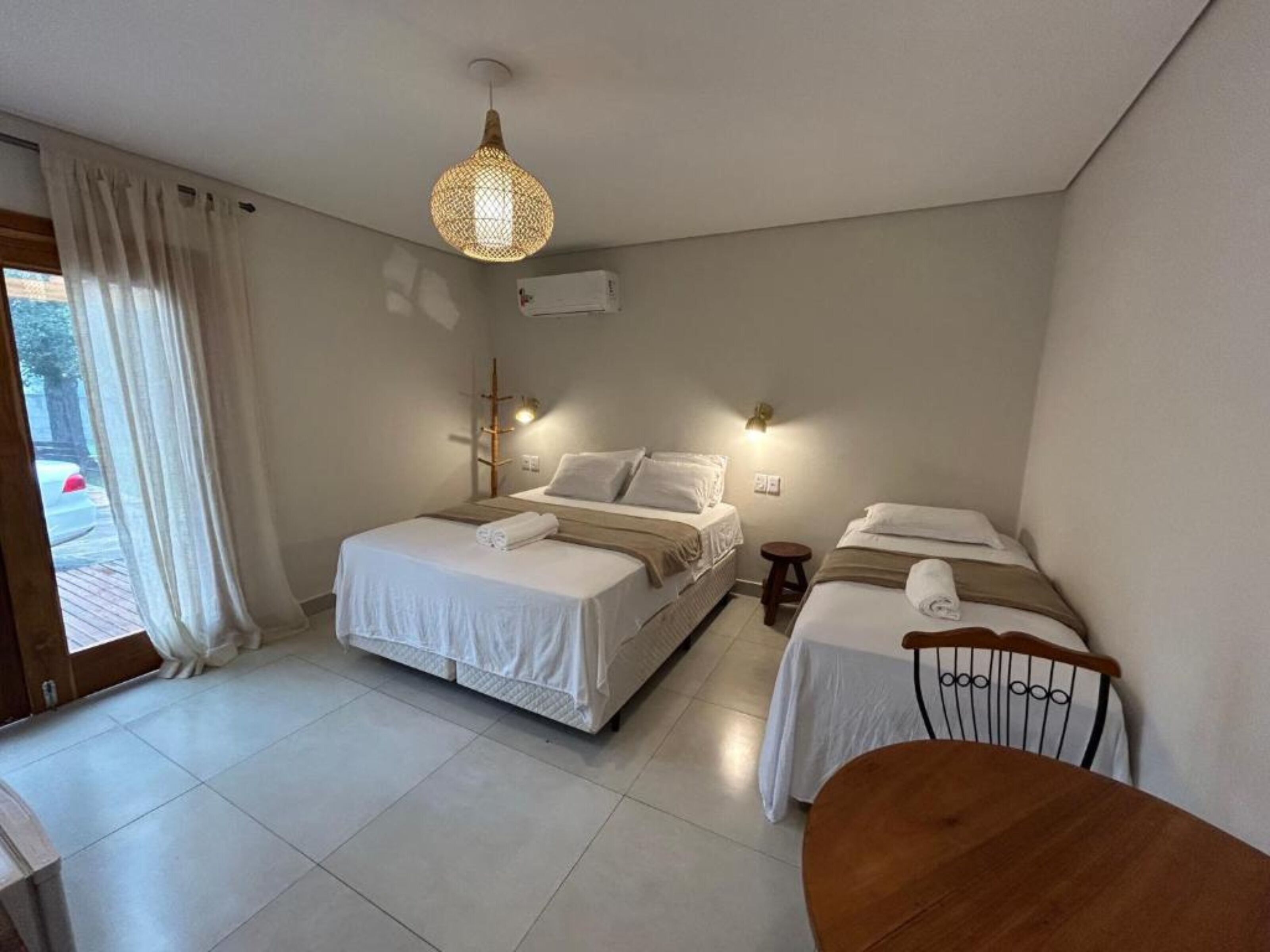 A hotel room with two beds, a wooden chair, a hanging light fixture, and a window with curtains.