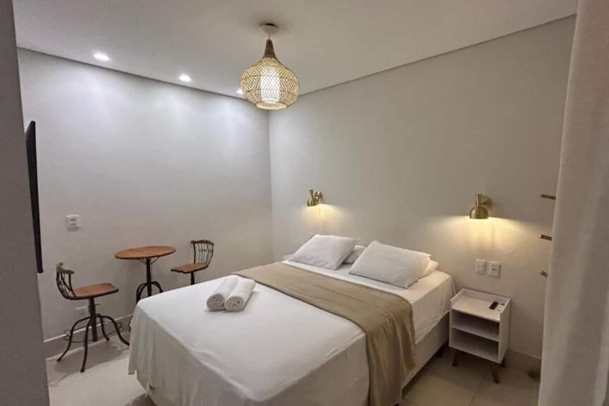 Una habitación de hotel con cama, mesitas de noche, lámpara, silla y una pequeña mesa.