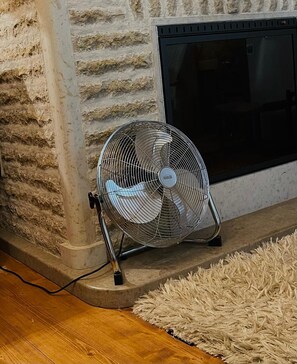 Un ventilatore a piantana vicino a un camino, una parete in pietra e un pavimento in legno.