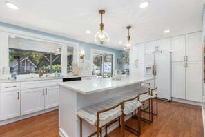 A kitchen with white cabinets, a large island with bar stools, pendant lighting, and a view through the windows. - Waterfront Getaway with Boat Slip (Ocean Pines)