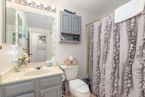A bathroom with a vanity, a toilet, a mirror with lights, a ruffled shower curtain, and a small plant on the wall. - Waterfront Getaway with Boat Slip (Ocean Pines)