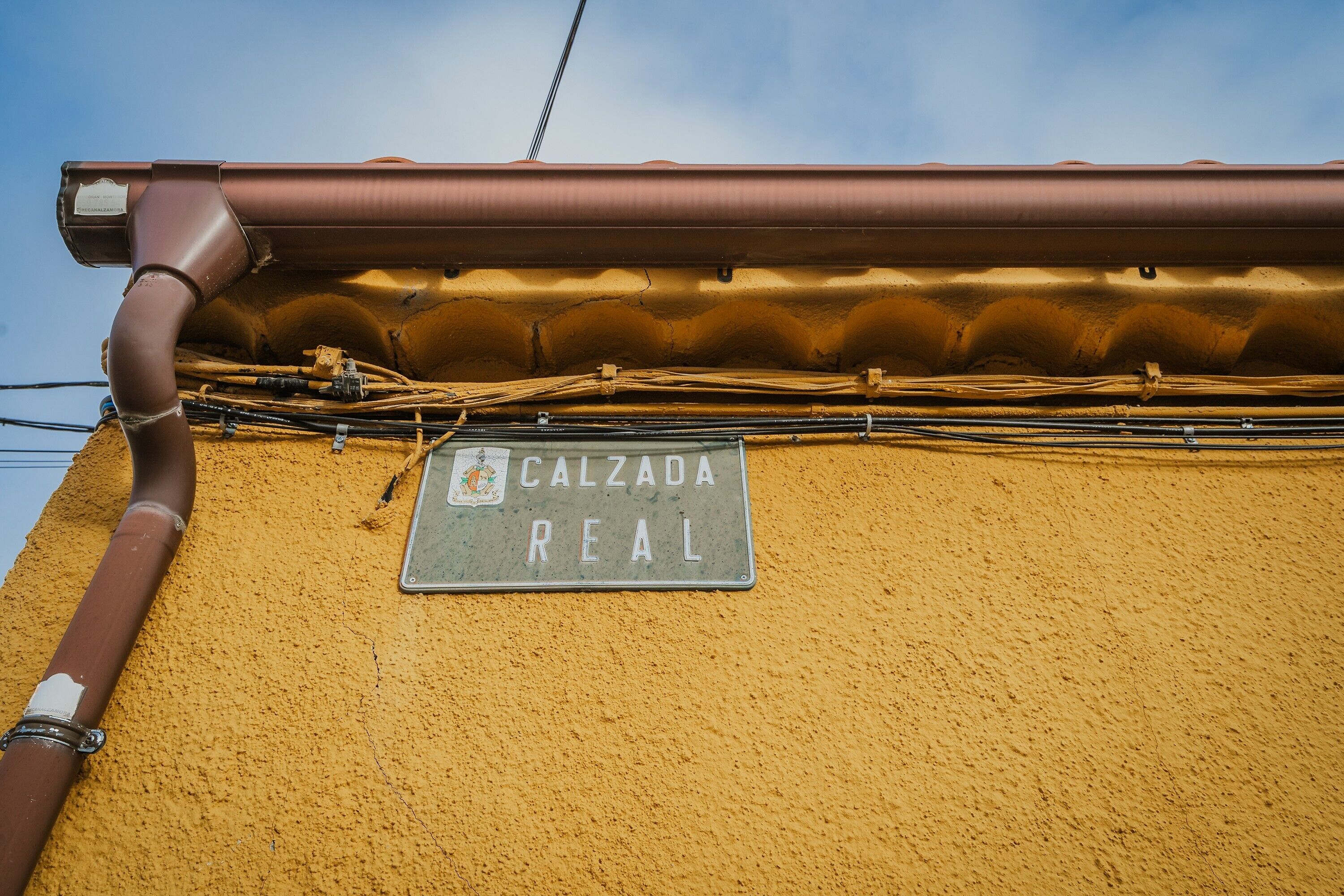 Ein Gebäudefassade mit einer braunen Regenrinne, einem Metallschild mit der Aufschrift 'CALZADA REAL' und einer strukturierten, gelben Wand.